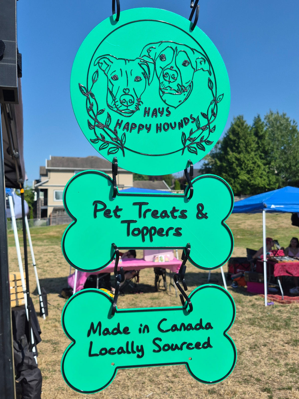 A bright turquoise green outdoor market sign hanging from a black metal stand. The sign is shaped like a dog bone and reads 'Pet Treats & Toppers' and 'Made in Canada Locally Sourced' in black lettering.