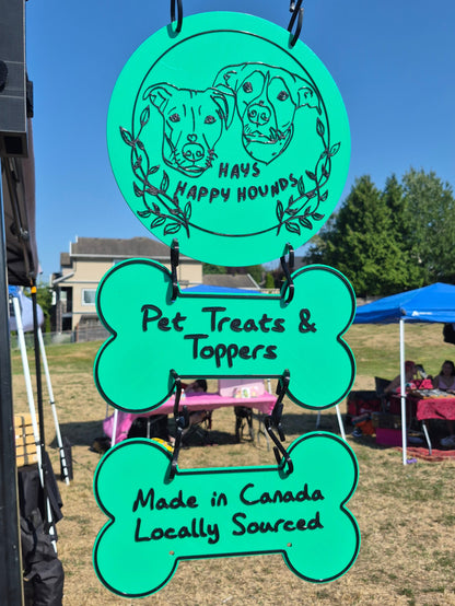 A bright turquoise green outdoor market sign hanging from a black metal stand. The sign is shaped like a dog bone and reads 'Pet Treats & Toppers' and 'Made in Canada Locally Sourced' in black lettering.